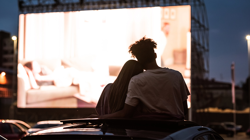 A couple with their arms around each other sit outside watching a drivethru film