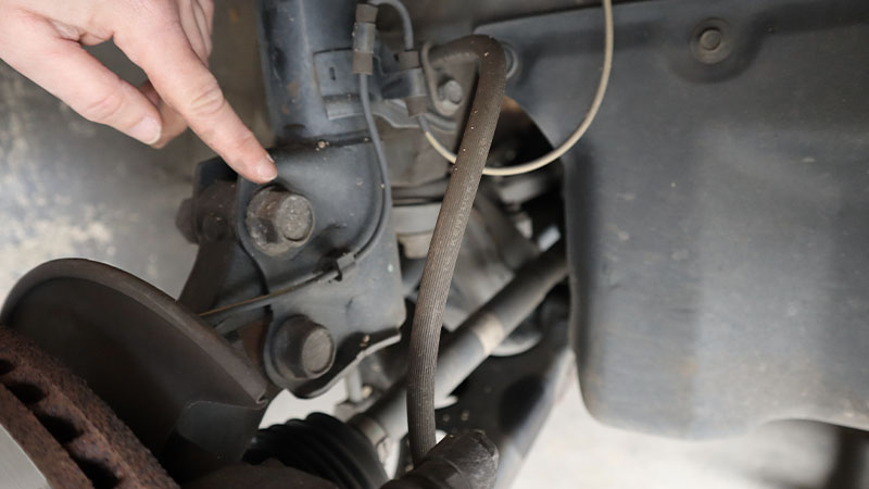 a hand points to one of two bolts that secure the steering knuckle/hub to the strut