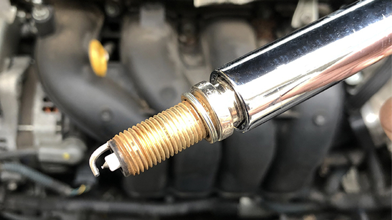 a close-up of an old spark plug inside the spark plug socket with a 4-cylinder engine in the background