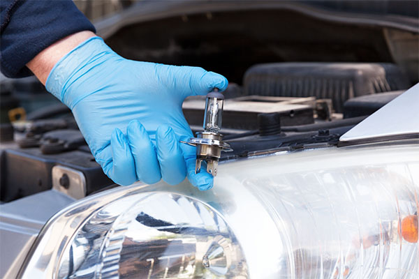 a gloved hand holding up a new headlight bulb in front of a vehicle with the hood up