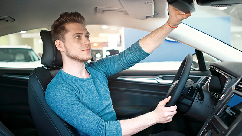 a young man sits in the driver's seat, holding the steering wheel with one hand and adjusting the rearview mirror with the other